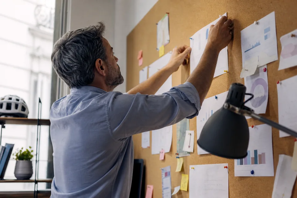 An agency wall with pinned printed report pages and sticky notes, a person reaching to pin another page