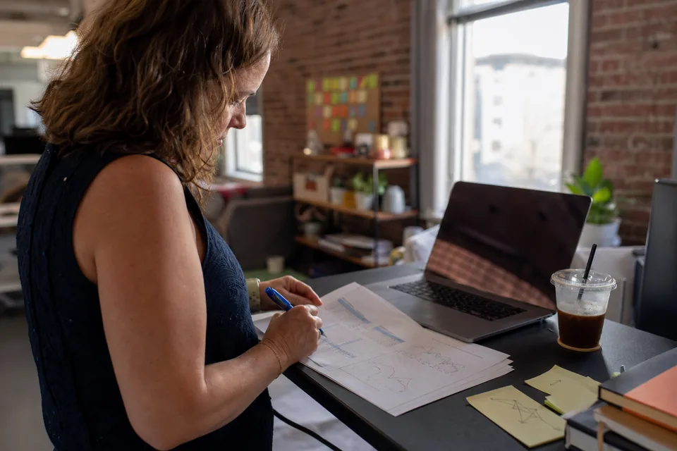 An analyst at a standing desk marking priorities with a pen on a printed domain report, laptop open beside it
