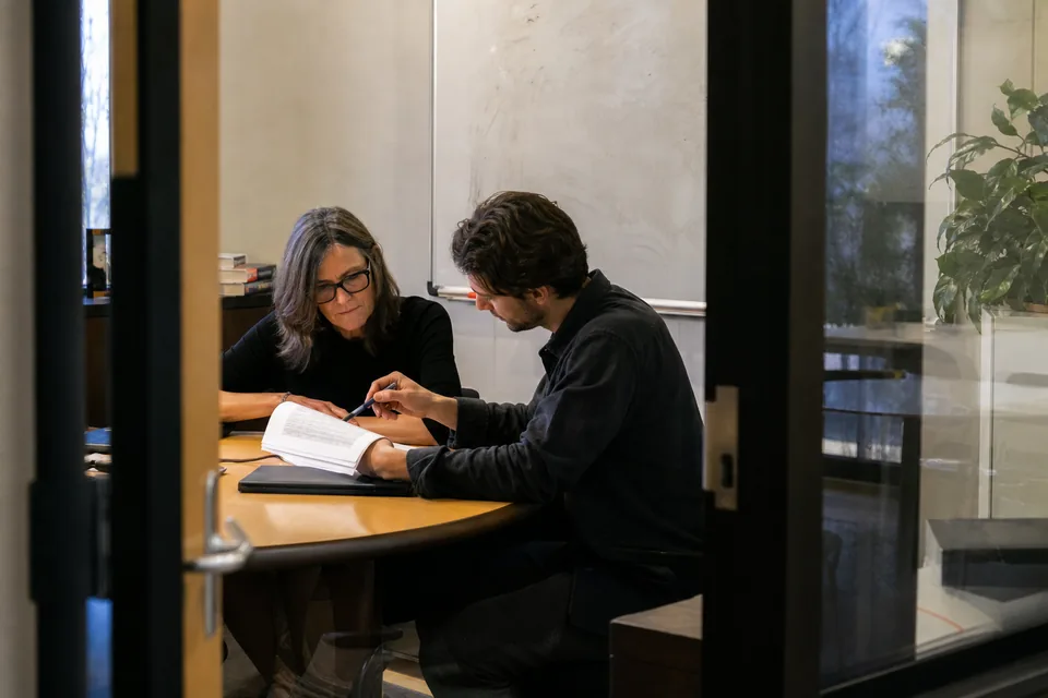 Two colleagues at a small meeting-room table going through a stapled printed domain report, one pointing with a pen